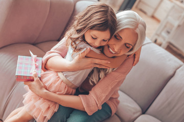 cheerful granddaughter hugging her grandmother with present in the hand at home