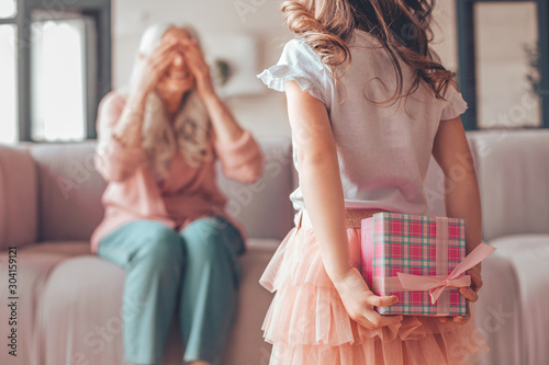 girl holding present box in the hands behind her back and making surprise for grandmother sitting on the couch
