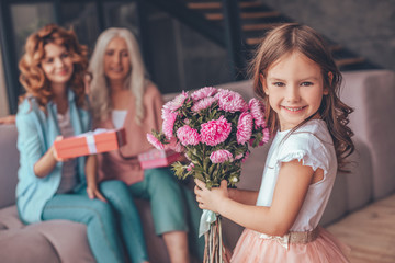 little girl holding bouquet of flowers and smiling at the camera while mother and grandmother sitting on the sofa