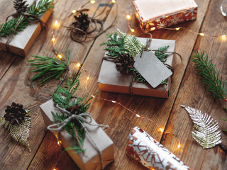 Christmas presents wrapped in craft paper with natural fir tree and thuja branches as decoration. Wooden table with light bulbs and hand made New Year gifts.