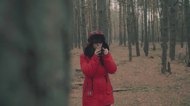 A portrait of the pretty woman with a cup of tea in woods. The woman in a jacket and a hat with earflaps is drinking a tea in a cold weather.