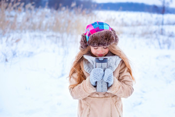 Fototapeta na wymiar Beautiful girl wearing beige jacket, colorful hat and grey knitted gloves drinks hot tea from thermal cup and walks in snowy park. Family winter vacation with kids in mountains on Christmas holiday