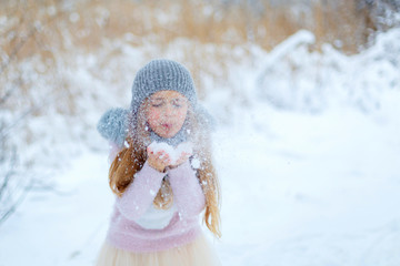 Pretty little girl wearing grey knitted hat and pink sweater walking in snowy park.  Cute child blowing snowflakes in winter forest. Family vacation with kid on christmas holidays. Blurred background