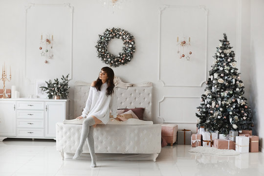 Beautiful young sexy woman in Christmas sweater posing at home in the morning. New year decorations. Sexy brunette model girl in knitted pullover and stockings posing over Christmas interior