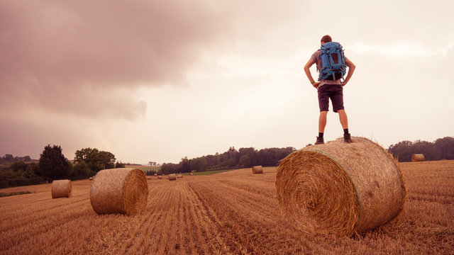 Fields, Ditzingen, Germany: Young man with backpack stands on bale of straw and overlooks the landscape while a thunderstorm is tearing in.