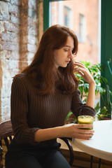 Beautiful serious stylish fashionable smart girl sits by the window in cafe and drinking healthy yellow smoothie or latte vegan. Charming thoughtful woman with long dark brown hair.