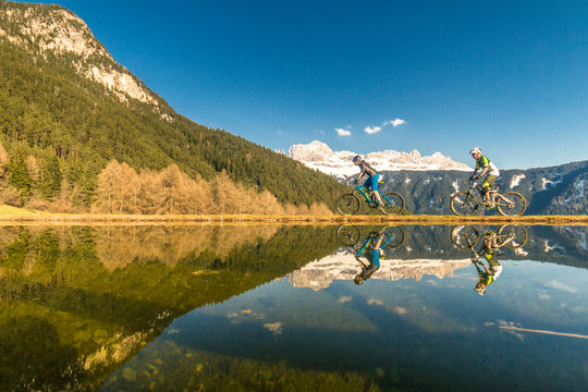 A male and female mountain biker riding along a lake in front of a mountain range in South Tirol, Italy.