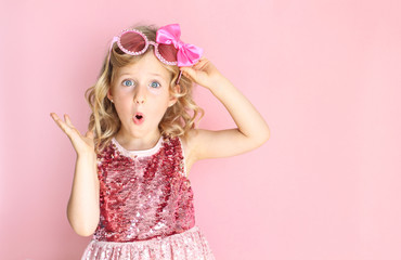 Portrait of a shocked girl wearing pink shiny dress and glasses. Little girl making a surprised expression. Bright and festive background. Party concept.