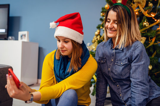 Two women friends female  mother and daughter or sisters sitting at home in front of the christmas tree taking photos selfies wearing santa hat using smart mobile phone video chat family new years eve