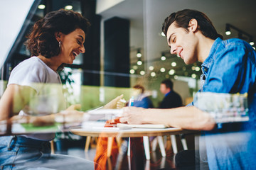 Side view of positive students having fun during exam preparation in cafe interior.Cheerful young man and woman laughing during collaboration on common studying task in modern coffee shop