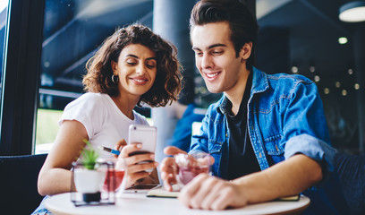 Positive young man and woman dressed in casual wear watching funny video on website on modern smartphone using 4G internet.Couple in love from millennial generation resting together in cafe