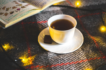 A cup of hot black tea with chocolate on a saucer on a warm and cozy plaid with Christmas lights