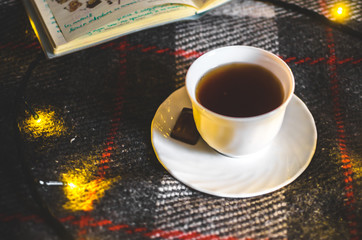 A cup of hot black tea with chocolate on a saucer on a warm and cozy plaid with Christmas lights
