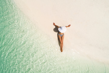 Summer holiday fashion concept - tanning girl wearing sun hat at the beach on a white sand shot from above.Top view from drone. Aerial view of slim woman sunbathing lying on a beach in Maldives. - obrazy, fototapety, plakaty