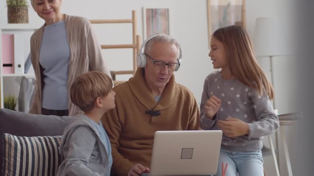 Medium shot of Caucasian boy and girl sitting on sofa with their granddad and helping him mastering laptop and headphones while aged woman watching them and then hugging