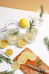 Jug with homemade lemonade, glasses with ice, lemons and rosemary on a white table.