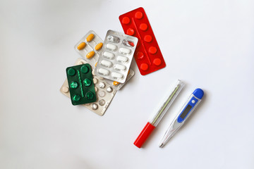 Heap of medical pills in white, green, red  and other colors. Pills in plastic package on a white background next to two types of thermometer-electric and mercury. Concept of healthcare and medicine 