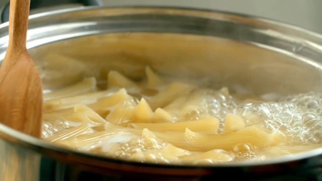 Penne pasta is cooked in boiling water, wooden spoon stirs pasta in slow motion side view