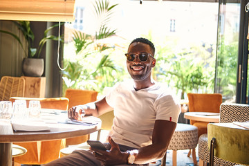 Happy smiling man in sunglasses sitting at cafe while chatting by mobile phone.