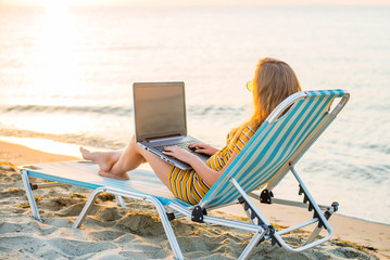 Fit young woman sitting on deck chair at sea view beach using laptop. Female freelance programmer in chaise-long lounge working coding surfing on notebook computer, blank screen. Remote work concept.