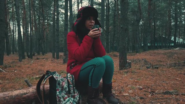 A pretty young woman in a jacket and a hat with earflaps is sitting on a log and drinking a tea in a cold weather in woods.