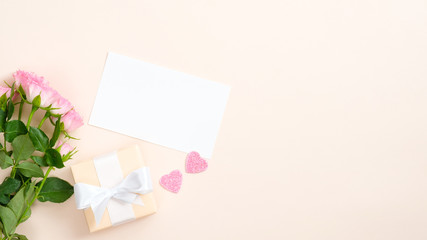 Gift box, blank paper card, roses, pink hearts on pastel beige background. Valentines Day, love, Mother day concept. Flat lay, top view, copy space.