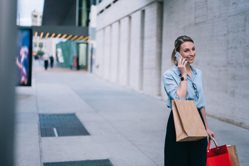 Cool cheerful hipster girl in stylish wear talking on smartphone in roaming, happy young woman calling on mobile phone device standing on street in modern city with paper bags during Black Friday