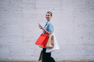 Half length portrait of cheerful female shopper with colorful paper bags in hands smiling at camera while strolling to next store with good offers of sales on Black Friday, happy woman near copy space