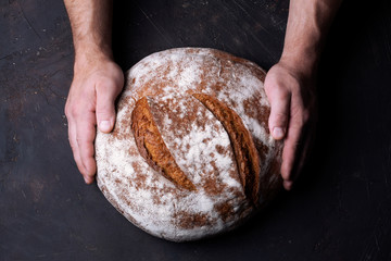 Baker holds big round rye bread in his hands on dark background top view