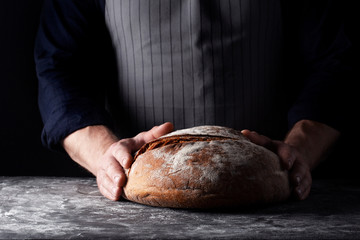 Baker holds big round rye bread in his hands on dark background