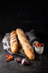 Fresh hot two baguettes, raw garlic and sun-dried tomatoes in olive oil on dark background.
