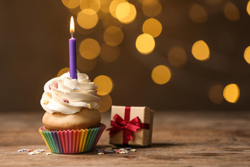 Birthday cupcake with candle and gift box on wooden table against blurred lights. Space for text