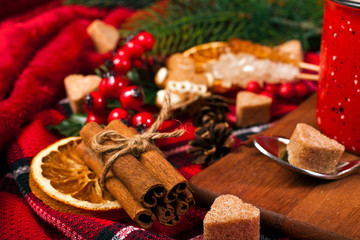 Christmas greeting card. Cacao drink in enamel cup with fir branches, dried orange slices, red berries, spices, heart shape sugar in spoon and gingerbread man on red fabric background.