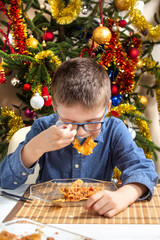 Chłopiec w okularach siedzi przy stole i ze smakiem zjada obiad. Bożonarodzeniowa choinka w tle. - obrazy, fototapety, plakaty