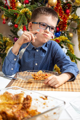 Chłopiec w okularach siedzi przy stole i ma buzię pełną jedzenia. Na talerzu leży lasagne a w tle pięknie ubrana choinka. Święta Bożego Narodzenia jedzenie. - obrazy, fototapety, plakaty