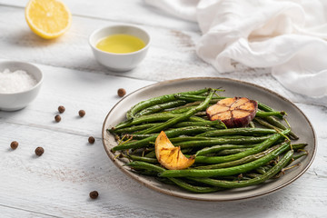 Plate with fried green beans, small bowls with olive oil and sea salt, white napkin, halved lemon and peppercorns on white wooden table. Healthy food concept