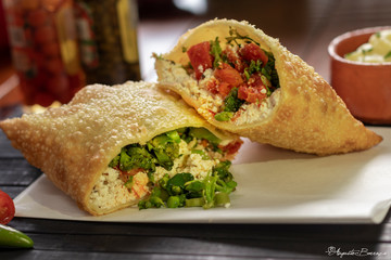 Pastel frito da culinária brasileira recheada com tomate seco e queijo ricota e legumes encima de um guardanapo  em uma mesa de restaurante com olho de maionese no fundo  e pimenta