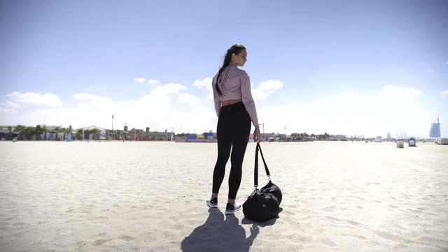 Beautiful young leggy fitness woman stands on the beach near the sea with a sports bag, starts walking. Slow motion