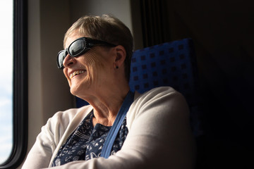 Obraz premium Smiling senior female tourist wearing sunglasses, looks out the window of a train while traveling in Munich, Germany.