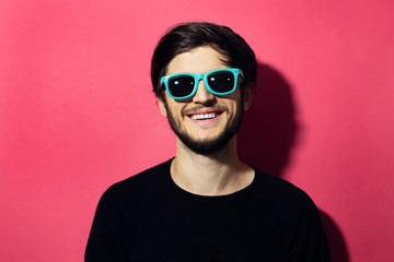 Portrait of young smiling man with cyan sunglasses on pink coral background.