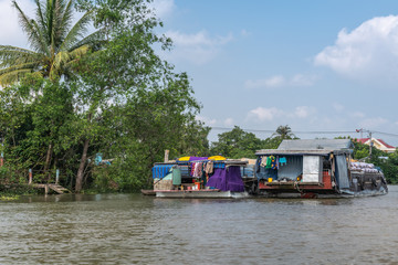 Fototapeta na wymiar Cai Be, Mekong Delta, Vietnam - March 13, 2019: Along Kinh 28 canal. 2 loaded up barges anchored at green foliage under blue cloudscape. Back of boat are family living quarters showing colors.