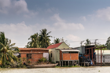 Cai Be, Mekong Delta, Vietnam - March 13, 2019: Along Kinh 28 canal. brown wooden sloop in front of red stone and yellow paint small farm house under blue cloudscape. Green foliage.