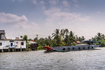 Cai Be, Mekong Delta, Vietnam - March 13, 2019: Along Kinh 28 canal. Modern gray motorized barge sailing on brown water under blue cloudscape. White house on stilts and green foliage.
