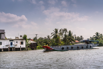 Fototapeta premium Cai Be, Mekong Delta, Vietnam - March 13, 2019: Along Kinh 28 canal. Modern gray motorized barge sailing on brown water under blue cloudscape. White house on stilts and green foliage.