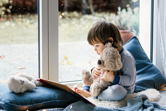 Cute little boy reading book hugging his teddy bear toy sitting cozy on pillows and knitted blanket  at window. Cozy home. Winter holidays lifestyle.