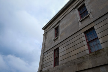 The exterior top corner of an old brick building with four windows. The wall is grey and textured. The sky is cloudy with some blue color. The roof is flat and the windows have red trim.