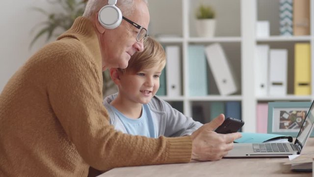 Medium shot of Caucasian boy showing elderly man sitting at desk how to use laptop, smartphone and headphones