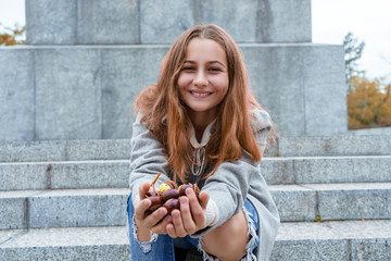 Beautiful girl is smiling and holds out hands with chestnuts. Teenager in gray coat outdoors in fall