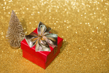 Red gift box with gold bow and little christmas tree on gold glitter surface with bokeh lights. 