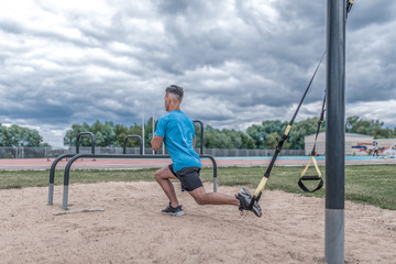 Young sportsman, male athlete, makes lunges with straps, loops for training legs, summer day city, fitness workout, active lifestyle modern youth, sportswear. Motivation life. Free space copy text.